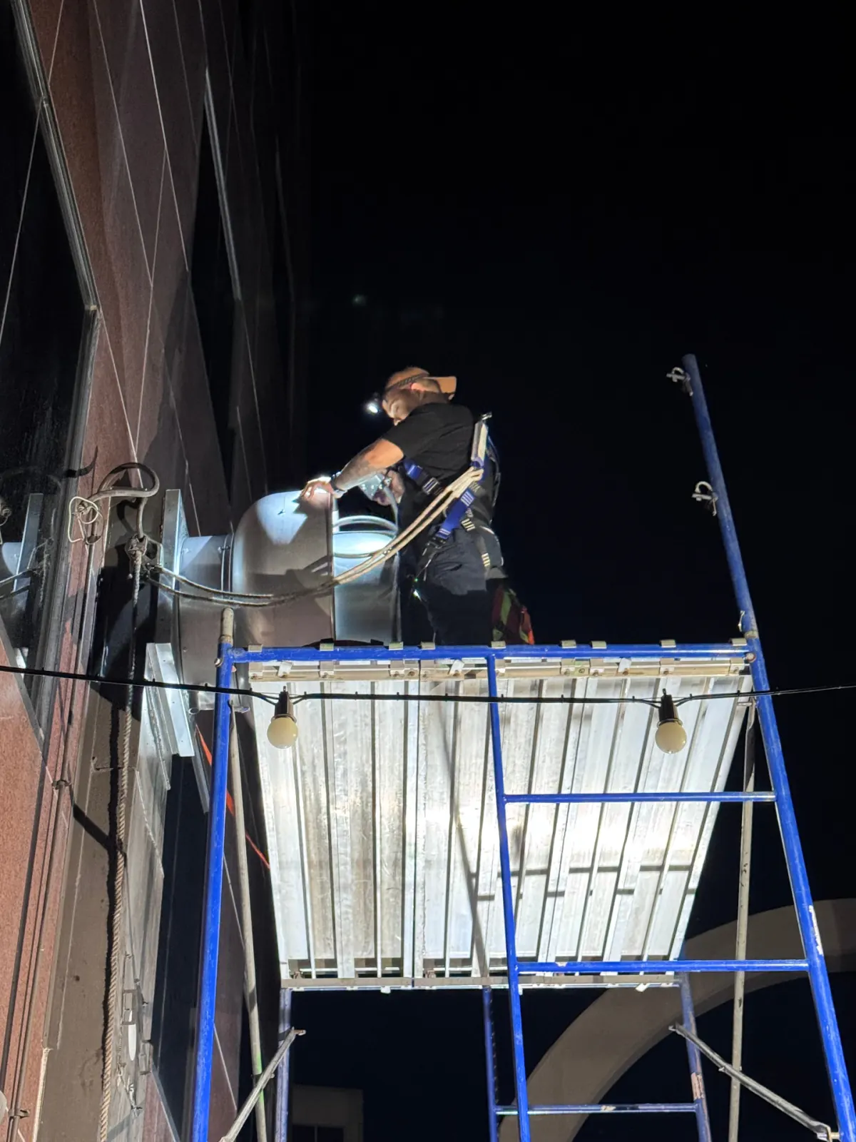 Technician servicing exterior exhaust fan from scaffold at night