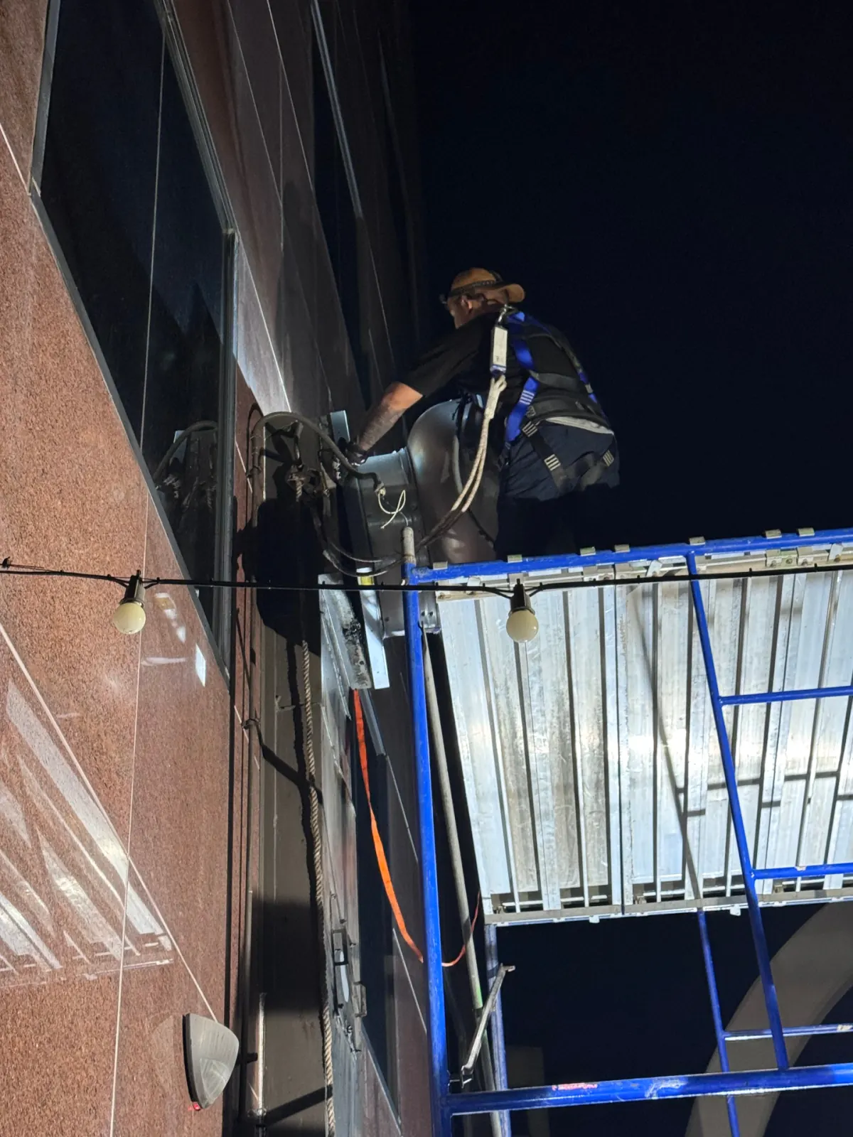 Technician harnessed on scaffold servicing rooftop exhaust fan at night