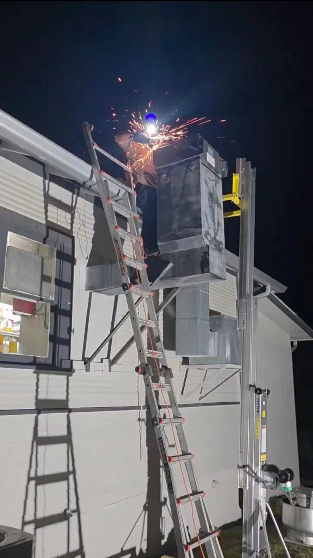 Welder repairing exterior exhaust duct with sparks flying at night
