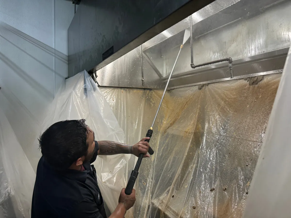 Technician pressure-washing inside commercial hood with containment sheeting