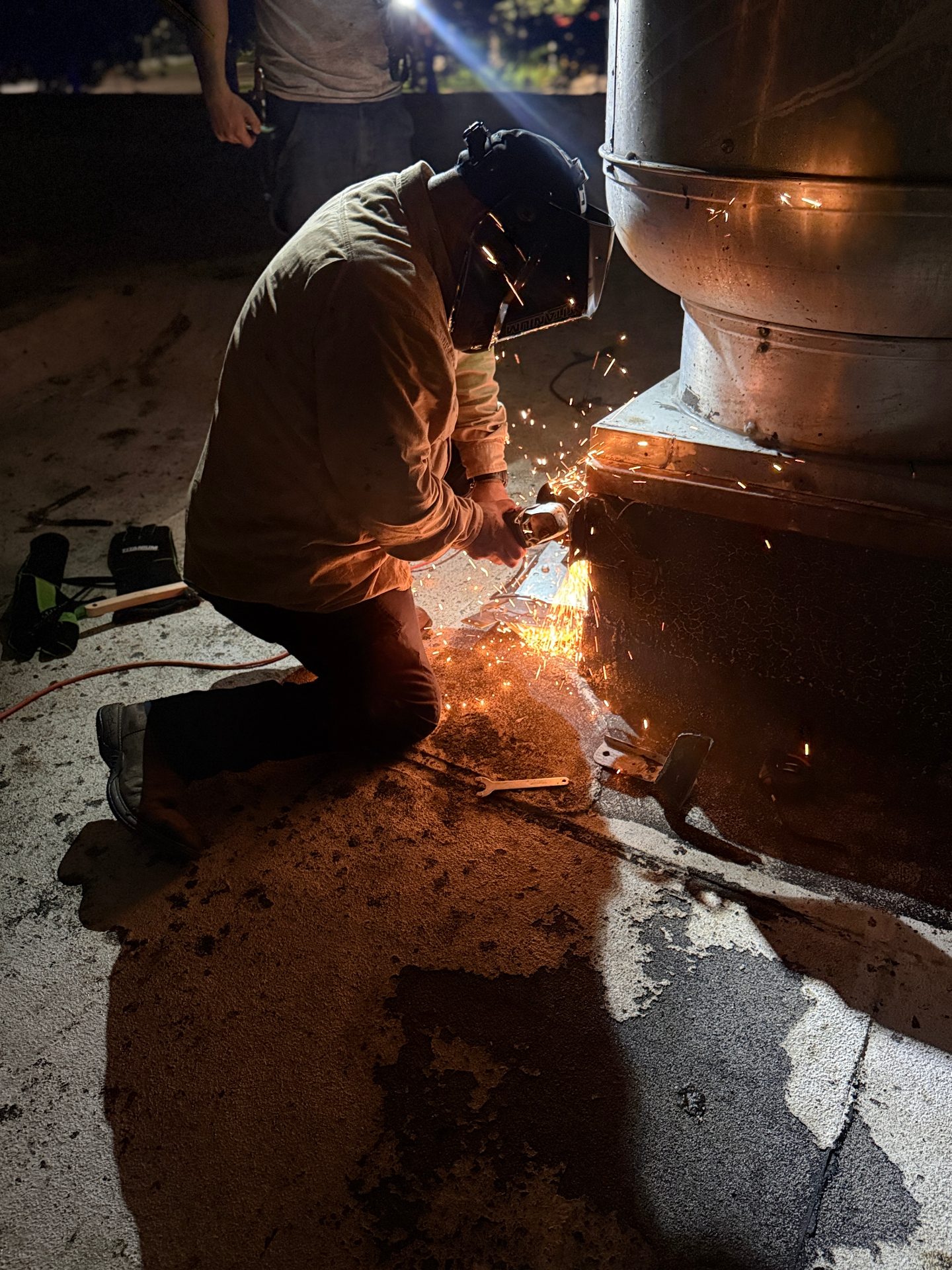 Welder in helmet cutting exterior exhaust fan base with sparks flying at night