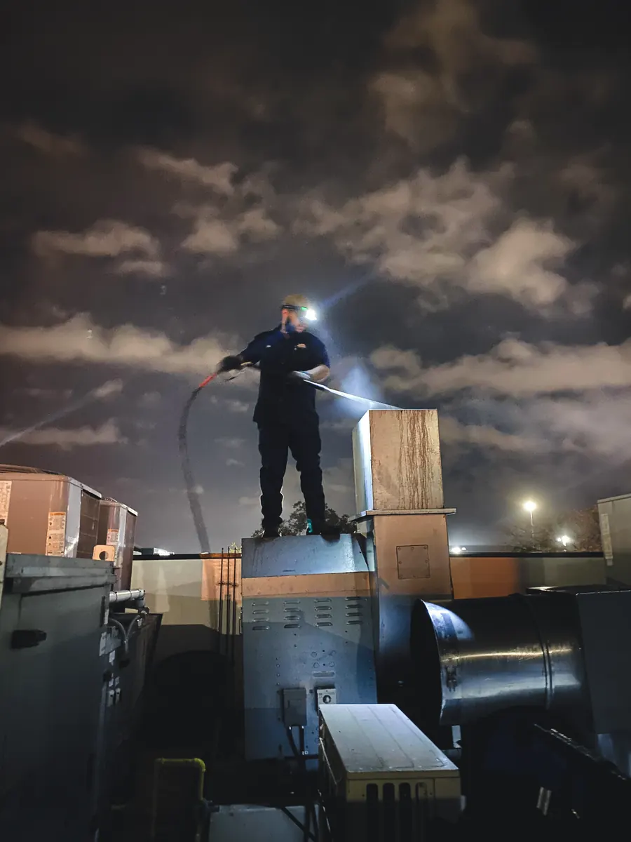 Technician pressure-washing rooftop ductwork at night